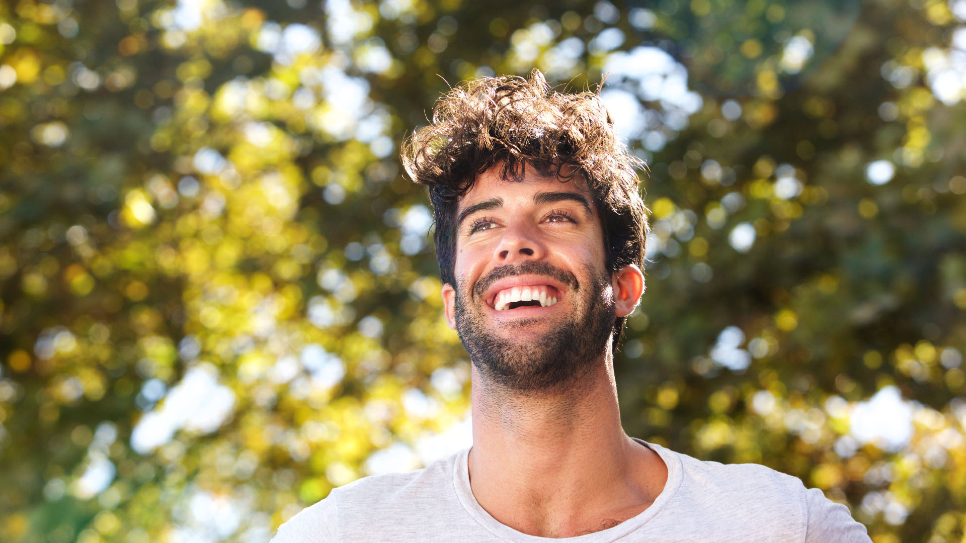 Young bearded man with curly hair smiling and looking upward outdoors, with blurred green trees behind him